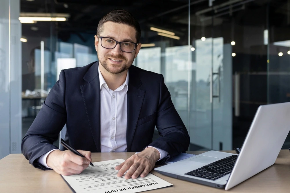 A Professional Man In Glasses Smiles At His Desk With A Laptop And Resume, Reflecting Business Culture And Ladders Job Search Success.