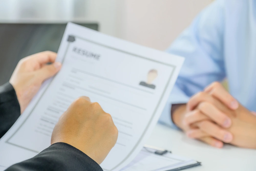 An Interviewer's Hand Holding A Resume With The Job Applicant In The Background